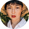 Woman with short hair wearing a wide-brimmed hat and a white shirt, surrounded by greenery, representing a fresh and vibrant approach to travel and passport services.