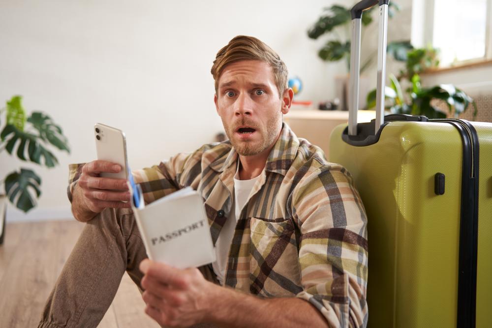 Man with a shocked expression sitting on the floor next to a suitcase, holding a smartphone and a passport, illustrating urgency in emergency travel preparations.